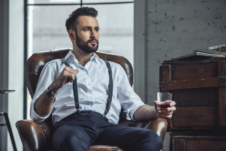 a handsome alcoholic business man is holding a glass of whiskey and looking away while sitting in leather armchair indoors