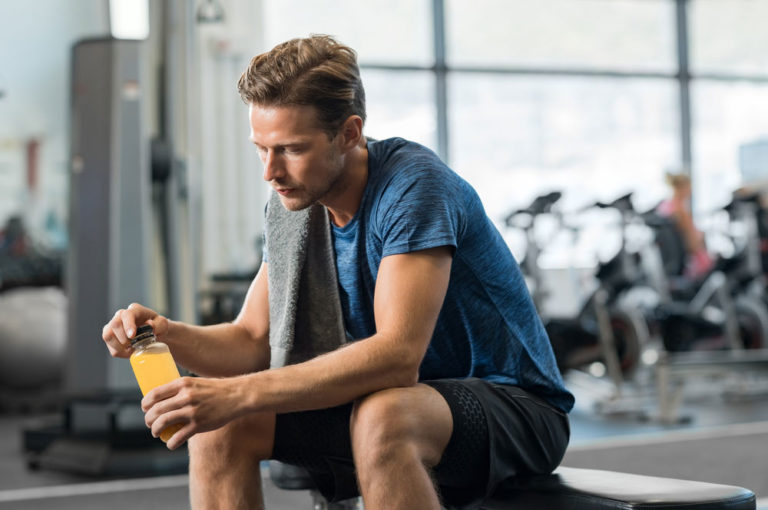 worried looking young man sitting in a gym holding a bottle of yellow liquid