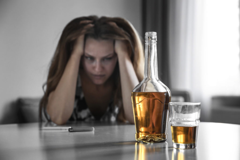 Glass with bottle of alcohol drink and drunk woman on background